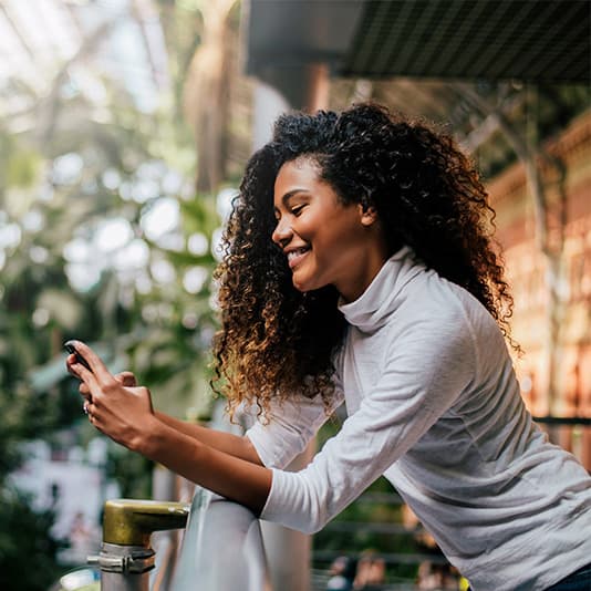 Smiling woman with curly hair wearing a white turtleneck, looking at her smartphone in a lush indoor setting.