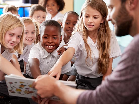 A diverse group of children in school uniforms eagerly pointing at a book held by a teacher, sitting together in a classroom setting.