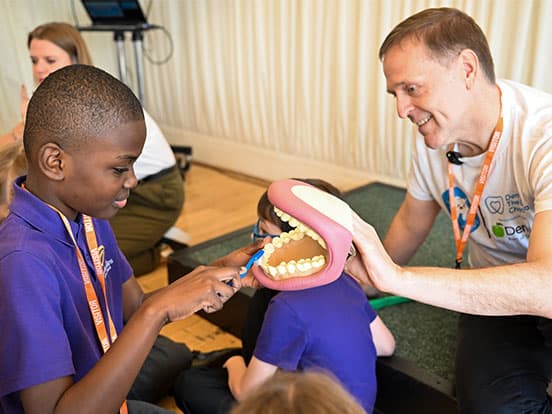 A boy in a purple shirt practices brushing on a large dental model, guided by Paul Schreier
