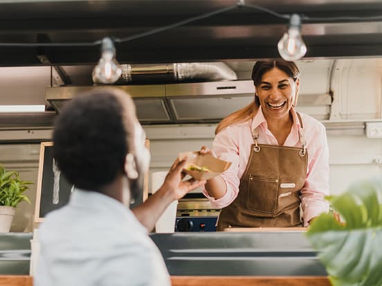 Smiling female vendor in a brown apron hands food to a customer through a food truck/window counter.