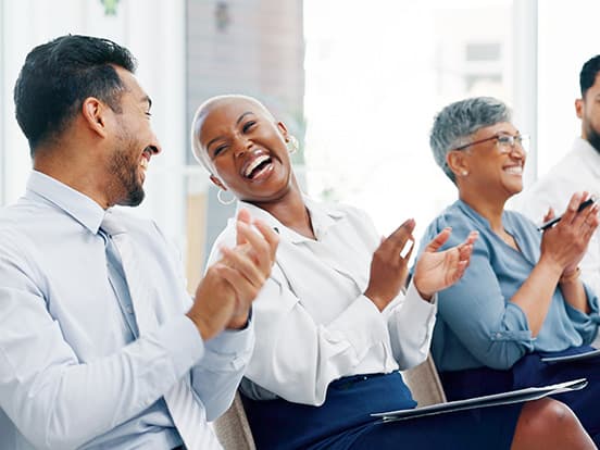 People sitting in a row, smiling and clapping, dressed in business attire, in a brightly lit room.