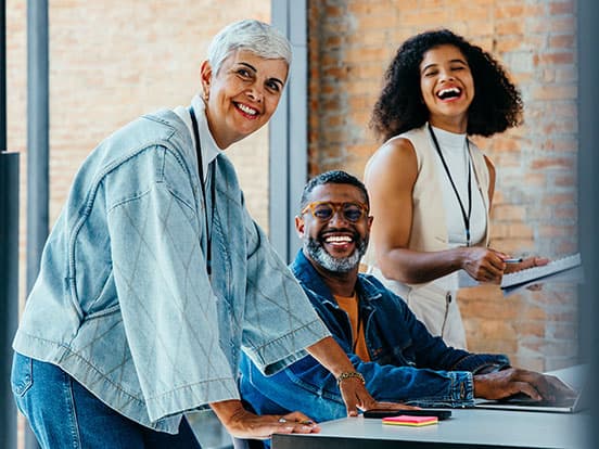 Three people smiling in a casual office setting, with one woman leaning on a table and two others seated, holding notebooks.