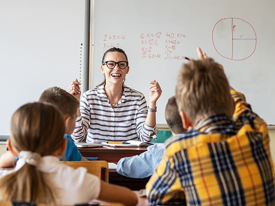 Smiling female teacher in glasses leads math lesson at whiteboard as elementary students raise hands.