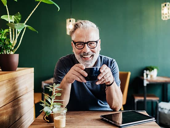 Smiling man with glasses holding a cup at a cafe table, surrounded by plants and an electronic tablet.