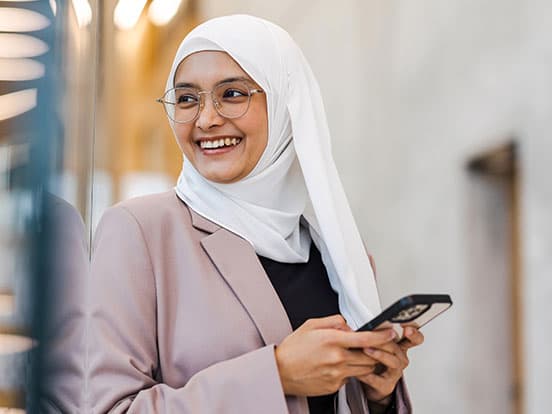 Smiling woman in a white hijab and glasses, holding a smartphone, wearing a mauve blazer in a modern, brightly lit setting.