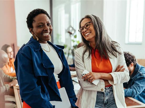 Two women smiling and chatting in an office, one holding a phone. People working in the background, with a bright window light.
