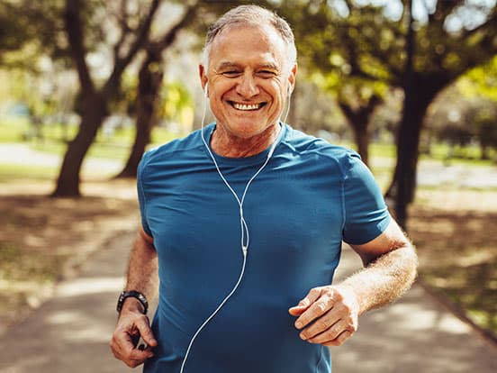 Smiling older man jogging on a tree-lined park path wearing a blue shirt and white earphones.
