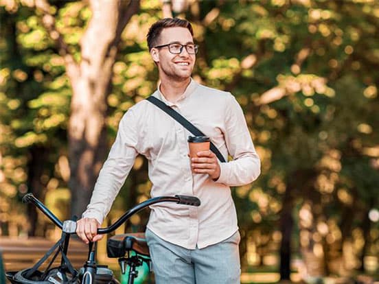 A man stands beside a bicycle in a park, holding a coffee cup, wearing glasses and a light-colored shirt, smiling and looking into the distance.
