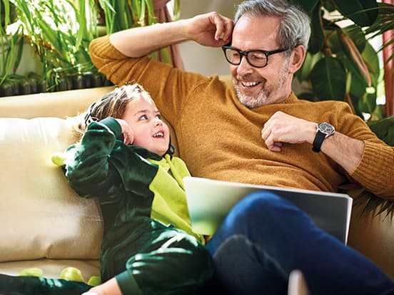 Man with a beard and glasses smiling at a child in a green costume on a couch, surrounded by plants, with a laptop nearby.