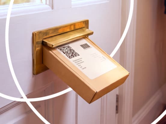 A package inserted halfway through a brass mail slot on a white door, with a visible label featuring a QR code and barcode.