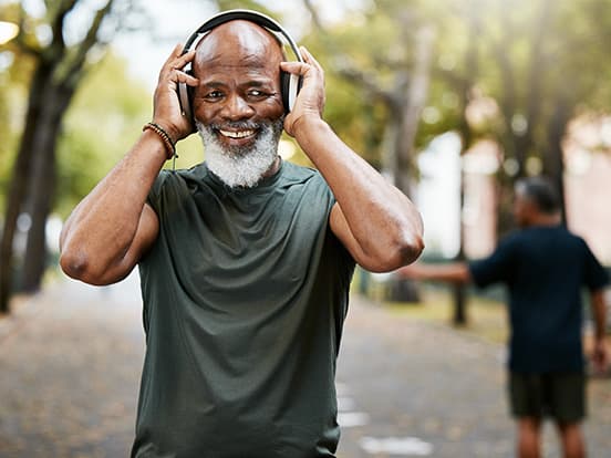 Elderly man smiling, wearing headphones and a green shirt, standing on a tree-lined path. Another person is blurred in the background.