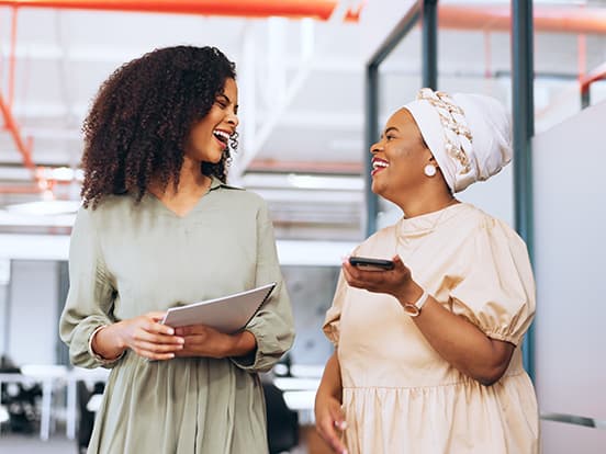 Two women in a modern office share a joyful conversation, one holding a tablet and the other a smartphone, both smiling brightly.