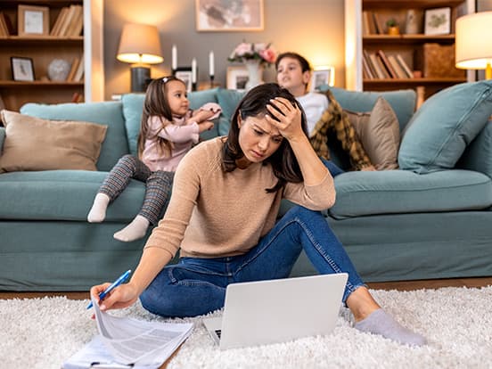 A woman sits on the floor, stressed, working on a laptop with papers, while two children play on a couch behind her in a cozy living room.