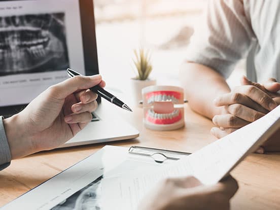 Two people in a dental office, discussing over documents. A dental model and X-ray on a laptop are visible on the desk.