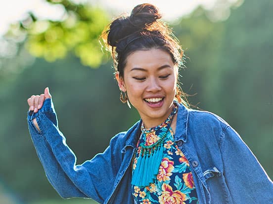 A woman with a joyful expression dances outdoors, wearing a floral dress and denim jacket, with trees in the background.