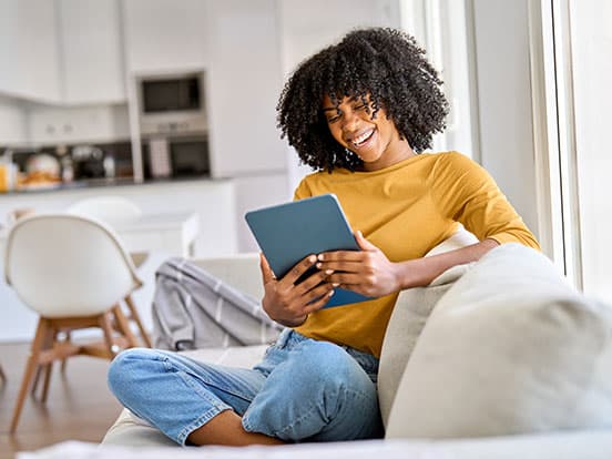 Woman sitting on a couch, smiling while reading a tablet in a bright, modern living room. She wears a yellow sweater and jeans.