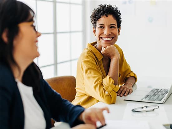 Two people sitting at a table; one smiles while looking at the other. A laptop and glasses are on the table. Bright, airy room in the background.