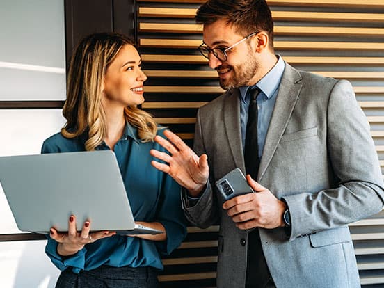 A man and woman in business attire smile while conversing; she holds a laptop, and he holds a smartphone.