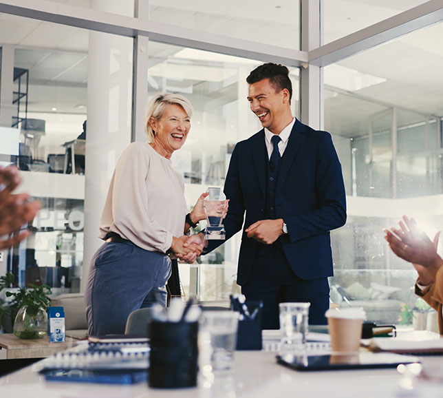 A woman and man, dressed in business attire, smile and shake hands while exchanging a glass plaque in a modern office setting.