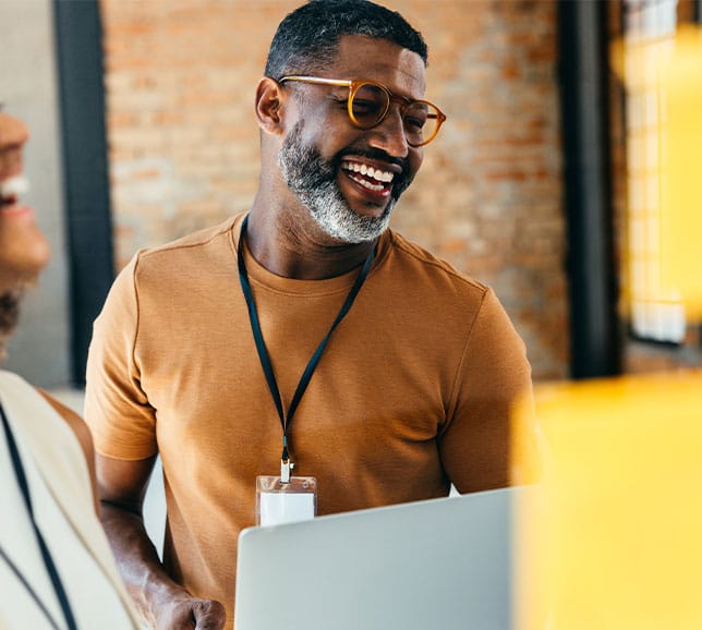 Smiling man with glasses and graying beard wearing a lanyard, leaning over a laptop in a sunlit office with exposed brick.
