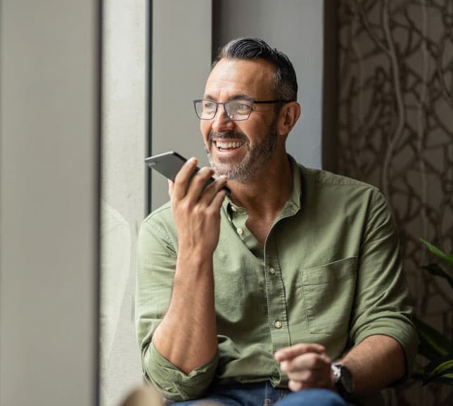 Smiling man with glasses in a green shirt, holding a smartphone near his mouth, seated by a window.