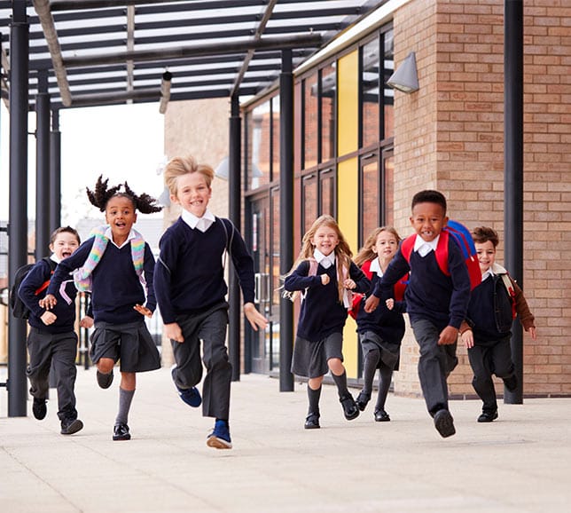 A group of cheerful children in school uniforms running outside a school building with backpacks on a sunny day.