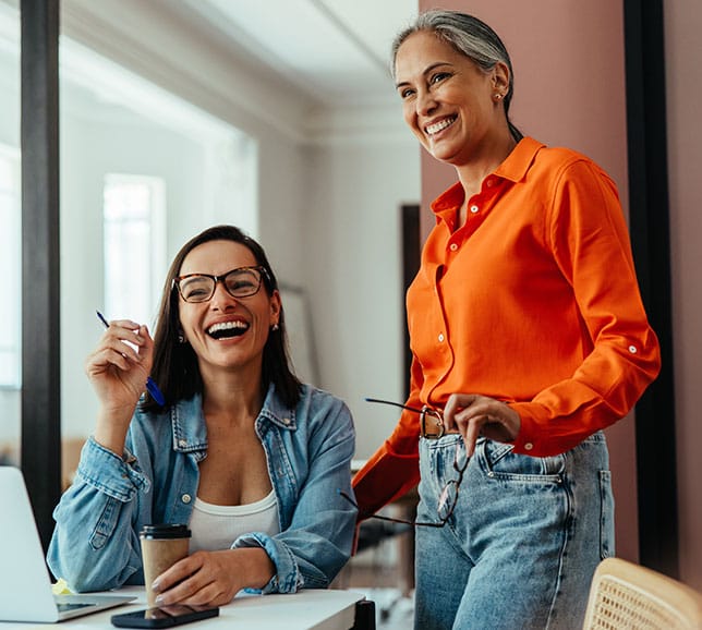 Two women smiling in an office environment. One is seated at a desk with a laptop and coffee, while the other stands, holding glasses.