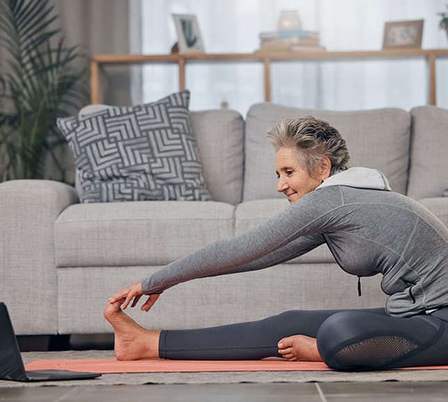 A woman in sports attire stretches on a yoga mat in a living room, reaching for her toes with one hand, smiling, and sitting on the floor.