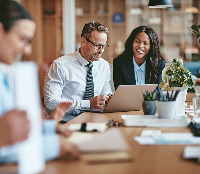 Two professionals at a desk, focused on a laptop, in a modern office setting. One is taking notes while the other looks on, both appearing collaborative.