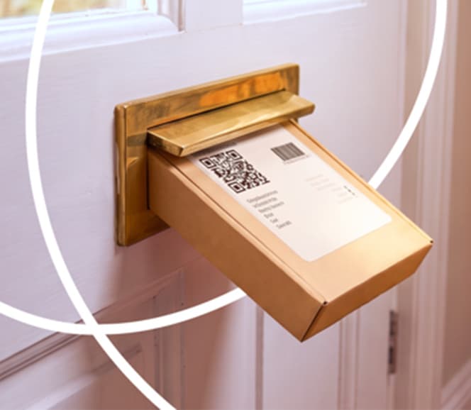 A small package with a QR code label is being pushed through a brass mail slot on a white door.