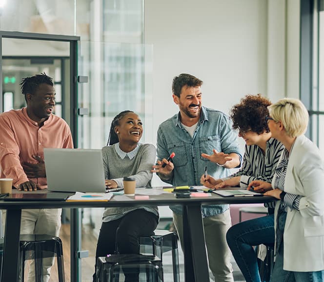 A group of five diverse colleagues laughing and collaborating around a desk with laptops and papers in a modern office environment.