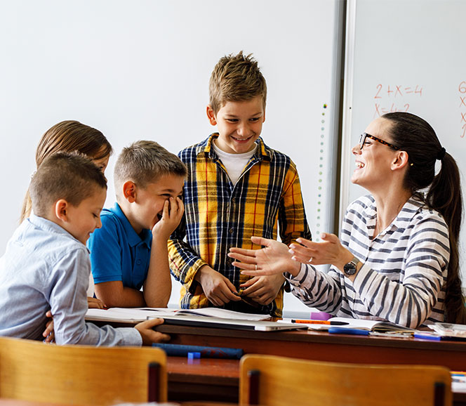Teacher and smiling students engage in a lively classroom discussion, with papers and books on the desk.