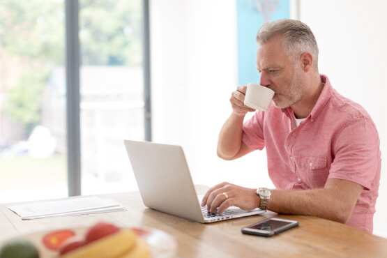 Older man sat at a kitchen table, drinking from a mug whilst on a laptop