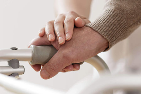 A caring hand placed on eldery person's hand using a walking frame