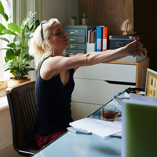 A woman sat at her desk at home stretching out forward taking a break from working